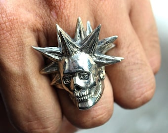 May include: A silver-colored ring featuring a skull design with spiky details. The skull has detailed facial features and is the focal point of the ring. The ring is a bold statement piece.