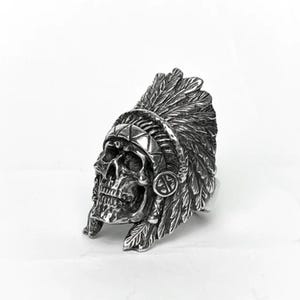 May include: A silver-colored ring featuring a skull wearing a Native American headdress. The headdress is detailed with feathers and geometric patterns. The ring has a detailed, textured appearance and is set against a white background.