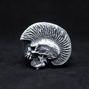 Op de afbeelding: Een zilveren ring met een gedetailleerd schedelontwerp met een mohawk. De schedel kijkt naar links en de mohawk bevindt zich aan de rechterkant van de schedel.