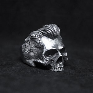 Könnte beinhalten: Ein silberner Totenkopfring mit einem detaillierten Design einer Pompadour-Frisur. Der Ring ist aus Metall und hat ein realistisches Totenkopfdesign.