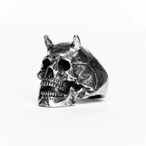 Puede incluir: Un anillo plateado con forma de calavera y pequeños cuernos. El anillo presenta un diseño detallado de calavera con dientes y cuencas de los ojos visibles. El anillo está sobre un fondo blanco, resaltando los detalles.