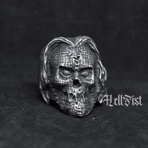 May include: A silver skull ring with a textured surface, resembling a stitched-up face. The ring is on a black background.