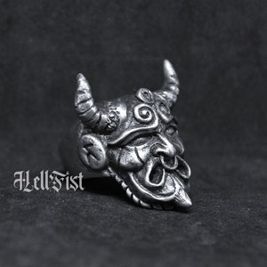 May include: A silver ring with a detailed demon mask design. The mask has horns, a wide mouth, and a textured surface.