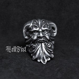 May include: A silver ring with a detailed design of a face with a beard and horns. The ring is on a black background.