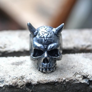 May include: A silver skull ring with horns and a pentagram on the forehead. The ring is on a gray concrete surface.