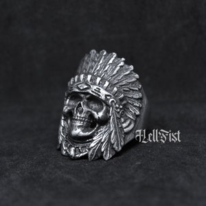 May include: A silver ring with a detailed skull wearing a headdress with feathers. The ring is engraved with the text "Hell Fist".
