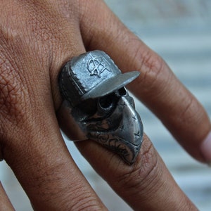 May include: A silver skull ring with a bandana and a baseball cap. The cap has an anarchy symbol on it.