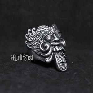 May include: A silver ring with a detailed design of a mythical creature's face. The creature has large, expressive eyes, a wide mouth, and a long, pointed tongue. The ring is a statement piece with intricate details.