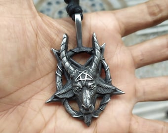 May include: A silver-toned pendant featuring a detailed depiction of a goat's head with horns, a pentagram, and a rope. The pendant is suspended from a black cord. The design is intricate and gothic in style.