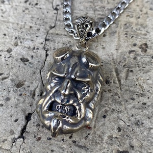 May include: A bronze-colored pendant necklace featuring a detailed, stylized face with horns and an open mouth. The pendant hangs from a silver-colored chain. The face has a distressed, aged appearance, suggesting a vintage or antique style.