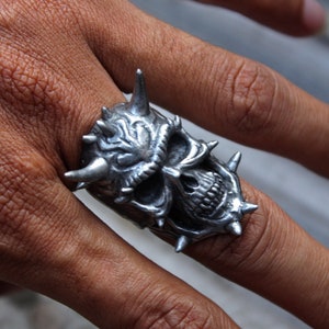 Puede incluir: Un anillo de calavera de plata con púas y cuernos. El anillo está detallado con una expresión amenazante y dientes afilados.
