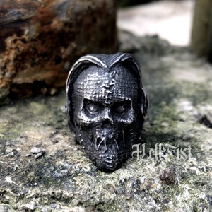 May include: A silver skull ring with a stitched-up face and a stitched-up mouth. The ring is on a gray, textured surface.