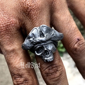Può includere: Un anello in argento a forma di teschio da pirata con un teschio dettagliato e un tricorno. L'anello è indossato su un dito.