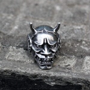 May include: A silver ring featuring a detailed design of a Japanese Oni mask.