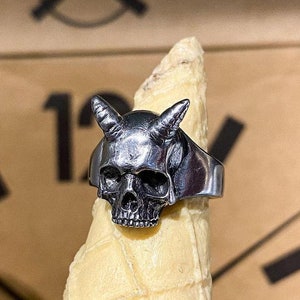 Puede incluir: Un anillo de plata con un diseño de calavera con cuernos. La calavera está detallada con cuencas de los ojos y una nariz.