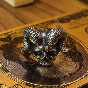 May include: A silver skull ring with horns. The ring is on a gold background.