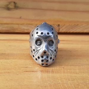 May include: A silver ring with a detailed design of a hockey mask. The mask has a series of holes and a raised design on the front.