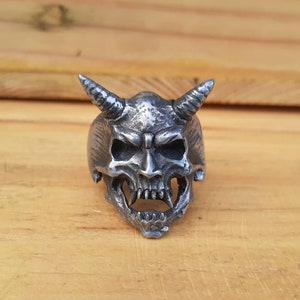 Puede incluir: Un anillo de plata con un diseño detallado de calavera de demonio. La calavera tiene cuernos, dientes afilados y una expresión amenazante.