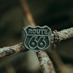 May include: A silver-colored ring featuring the Route 66 logo. The ring has a shield-shaped design with the words "ROUTE" above the number "66". The ring is set against a blurred, natural background.