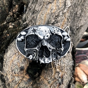 May include: A silver skull belt buckle with a black background. The skull has a cracked and weathered look, with a star on its forehead.