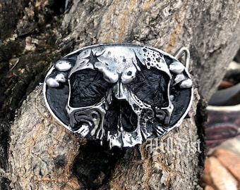 May include: A silver skull belt buckle with a black background. The skull has a cracked and weathered look, with a star on its forehead.