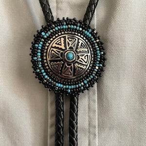 May include: A black leather bolo tie with a round turquoise and silver bead pendant. The pendant features a geometric design with a turquoise stone in the centre.