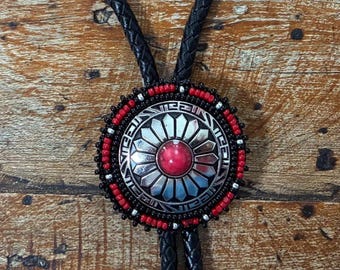 May include: A bolo tie with a circular, silver-toned pendant featuring a red center stone and intricate geometric designs. The pendant is bordered by red and black beads, and hangs from a black braided leather cord.