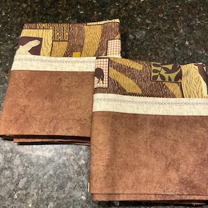 May include: Two folded brown fabric pieces with a textured appearance. The top section features a patterned design with brown, tan, and cream colors, including animal and leaf motifs. The fabric is likely used for home decor or crafting.