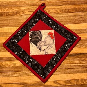 May include: A diamond-shaped fabric pot holder featuring a central square panel with a rooster illustration. The rooster has black tail feathers and a red comb. The panel is framed by a solid red border and an outer black border with white floral patterns.