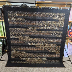 May include: A large, handmade quilt with a black border and a patchwork design. The quilt features horizontal bands of various patterned fabrics in shades of black, brown, and gold. The quilt is displayed on a black metal stand.