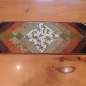 May include: A brown, green, and white quilted table runner with a geometric pattern. The runner is made of fabric with a pine cone and branch design.
