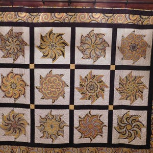 May include: A quilt with a black and white background and a repeating pattern of gold and brown swirls. The quilt is made up of 12 squares, each with a different swirl pattern.