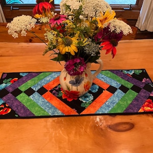 May include: A colorful floral arrangement in a decorative pitcher sits on a handmade table runner. The runner features a black background with a geometric pattern of purple, green, blue, and orange stripes. The table is made of wood.