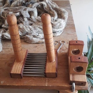 May include: A wooden wool carder with two handles and a metal comb. The carder is used to blend and prepare wool for spinning. A skein of gray wool is in the background.