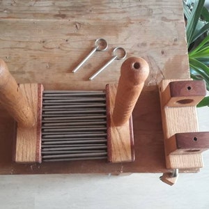 May include: A wooden wool carder with two handles and a metal comb. The carder is made of light brown wood with dark brown accents. The metal comb has many fine teeth.