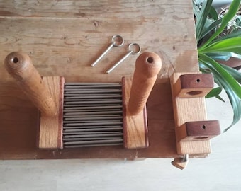 May include: A wooden wool carder with two handles and a metal comb. The carder is made of light brown wood with dark brown accents. The metal comb has many fine teeth.
