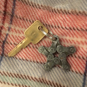 May include: A gold key with a silver star-shaped keychain made from bike chain links. The keychain is on a red and white plaid fabric.