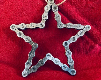 May include: A silver star-shaped ornament made from a bicycle chain. The ornament is hanging from a piece of twine against a red background.