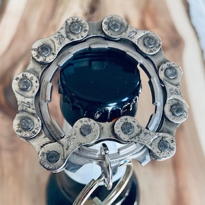 May include: A silver bottle opener crafted from a bicycle chain, designed to remove bottle caps. The opener is circular, with the chain links forming the outer edge. A key ring is attached. The opener is on top of a dark bottle.