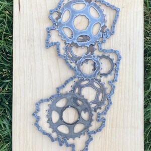 Puede incluir: Una escultura de metal del estado de Nueva Jersey hecha con cadenas y engranajes de bicicleta. La escultura est&aacute; montada sobre un fondo de madera clara.