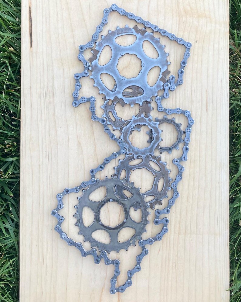 Puede incluir: Una escultura de metal del estado de Nueva Jersey hecha con cadenas y engranajes de bicicleta. La escultura est&aacute; montada sobre un fondo de madera clara.