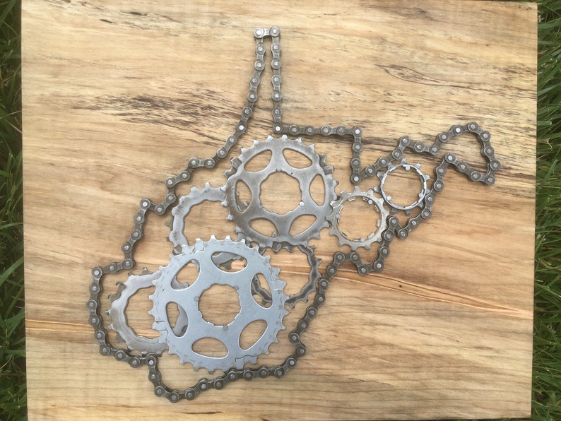 Puede incluir: Una pieza de arte met&aacute;lico con la forma del estado de Virginia Occidental, hecha de cadenas y engranajes de bicicleta. La pieza est&aacute; montada sobre un fondo de madera.
