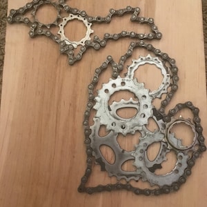 Puede incluir: Una pieza de arte de metal con la forma del estado de Michigan, hecha de cadenas y engranajes de bicicleta. La pieza est&aacute; montada en una tabla de madera.