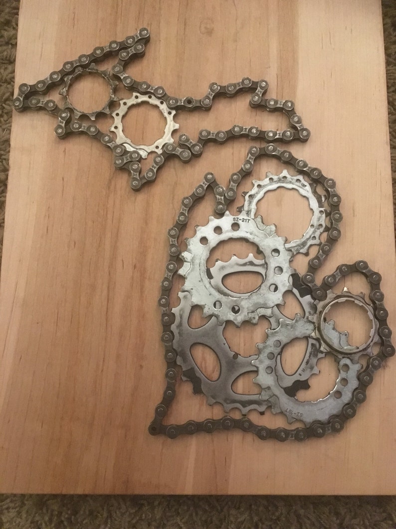 Puede incluir: Una pieza de arte de metal con la forma del estado de Michigan, hecha de cadenas y engranajes de bicicleta. La pieza est&aacute; montada en una tabla de madera.