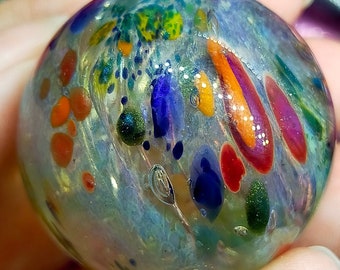Pode incluir: Uma bola de gude de vidro transparente com desenhos coloridos embutidos. A bola de gude apresenta uma mistura de pontos e listras laranja, vermelhas, azuis, verdes e amarelas. O vidro tem uma qualidade ligeiramente iridescente, com pequenas bolhas de ar visíveis.