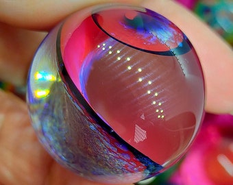 Pode incluir: Uma bola de gude de vidro transparente com um design vibrante. A bola de gude apresenta padrões em espiral de vermelho, rosa e azul, com uma linha de destaque preta. O vidro reflete a luz, criando um efeito brilhante. A bola de gude é segurada em uma mão.