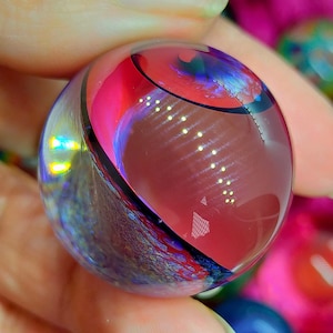 Könnte beinhalten: Eine transparente Glasmurmel mit einem lebendigen Design. Die Murmel weist wirbelnde Muster aus Rot, Rosa und Blau mit einer schwarzen Akzentlinie auf. Das Glas reflektiert das Licht und erzeugt einen schimmernden Effekt. Die Murmel wird in einer Hand gehalten.