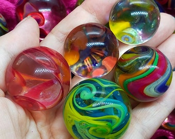 May include: A collection of spherical glass marbles featuring various internal designs, including swirling patterns of red, blue, green, yellow, and orange colors. The marbles have a smooth, polished surface and are translucent, allowing light to pass through the glass.