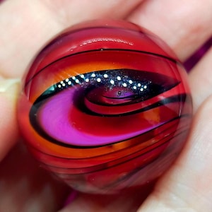 May include: A spherical glass marble featuring a swirling pattern of deep red, vibrant orange, and magenta hues. The surface is accented with thin, dark black lines that curve around the sphere, and a central band of tiny, reflective white dots.