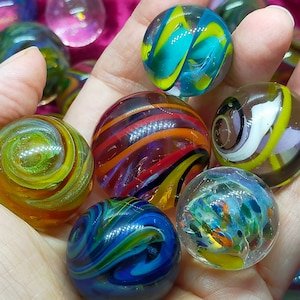 Può includere: Una collezione di biglie di vetro colorate con motivi a spirale. Le biglie presentano vari colori, tra cui blu, giallo, rosso e verde. Alcune biglie hanno disegni a strisce, mentre altre hanno motivi astratti. Le biglie sono tenute in una mano.